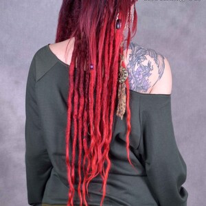 May include: A woman wearing a dark green off-the-shoulder sweatshirt with long red dreadlocks. The sweatshirt has a ribbed olive green band around the bottom. The woman is wearing black pants with a red, yellow, and green stripe down the side.