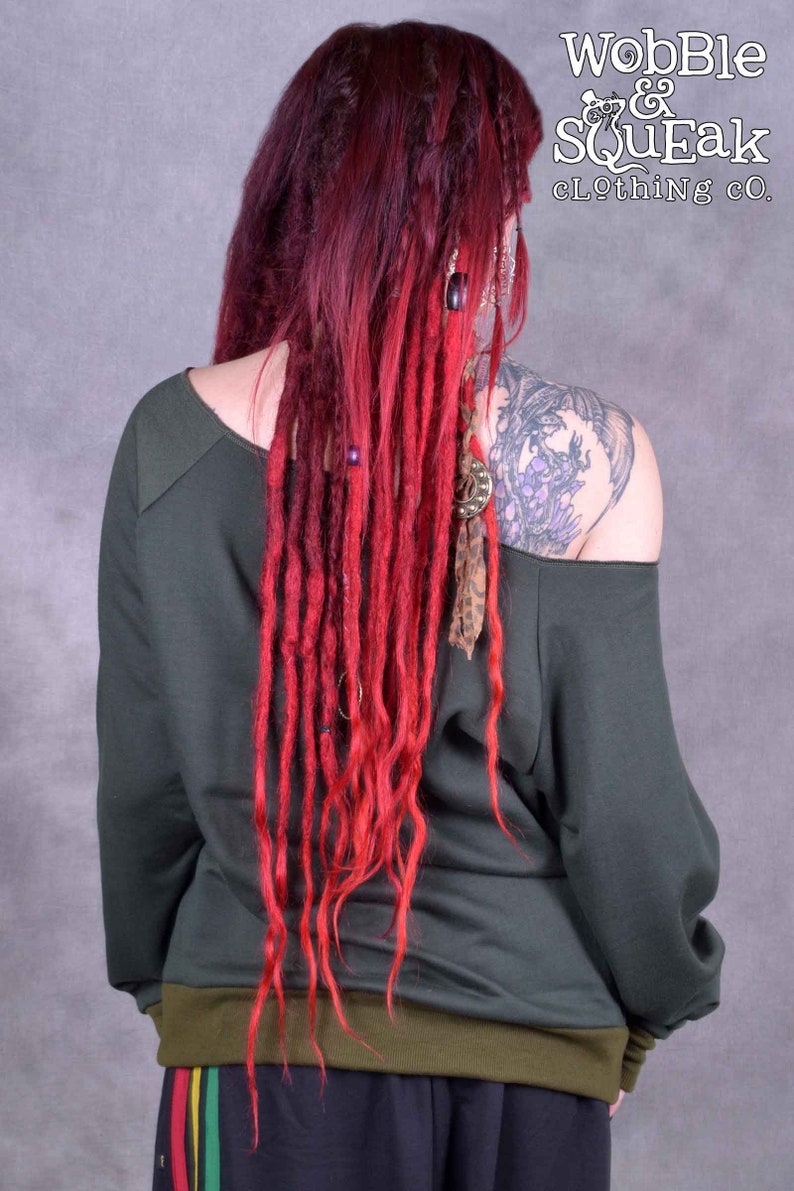 May include: A woman wearing a dark green off-the-shoulder sweatshirt with long red dreadlocks. The sweatshirt has a ribbed olive green band around the bottom. The woman is wearing black pants with a red, yellow, and green stripe down the side.