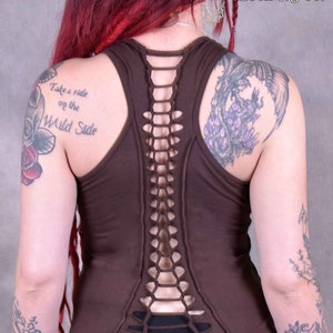 Peut inclure: Top sans manches marron avec un détail de laçage dans le dos. Le haut est fait d'un tissu doux et confortable et a une coupe décontractée. Le mannequin porte le haut avec un pantalon noir et a les cheveux roux. Le texte "Take a ride on the Wild Side" est visible sur l'épaule gauche du mannequin.