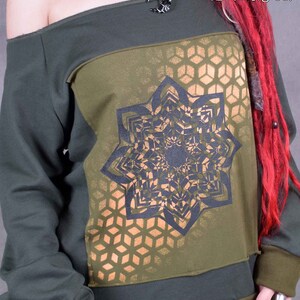 May include: A green off-the-shoulder sweatshirt with a black and gold geometric design. The design features a large mandala-like pattern with a honeycomb background. The text "Wobble & Squeak clothing co." is printed on the top right corner of the sweatshirt.