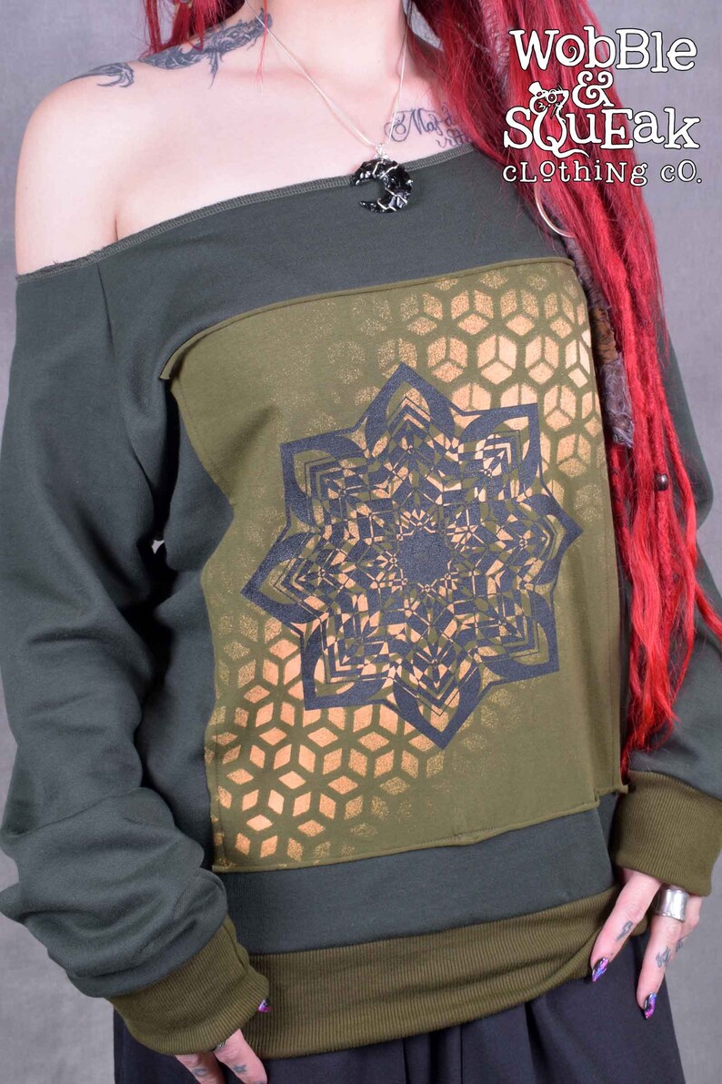 May include: A green off-the-shoulder sweatshirt with a black and gold geometric design. The design features a large mandala-like pattern with a honeycomb background. The text "Wobble & Squeak clothing co." is printed on the top right corner of the sweatshirt.