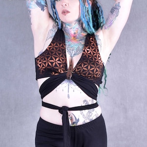 May include: A black halter top with a copper-coloured geometric pattern. The top has a wrap-around design and ties at the waist. The model is wearing black trousers.