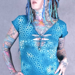 May include: A teal blue t-shirt with a white geometric pattern. The shirt has a deep v-neck and a lace-up detail at the front. The model is wearing the shirt with black trousers.