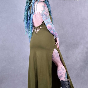 A woman wearing a long, olive green maxi dress with a lace-up back and a side slit. The dress is made of a soft, stretchy fabric and has a relaxed fit. The woman is wearing black boots with buckles.