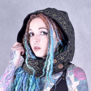Peut inclure: Cagoule noire avec motif géométrique doré et boutons en bois. La cagoule est doublée de fausse fourrure noire. Le modèle porte la cagoule et a des dreadlocks bleus et bruns.
