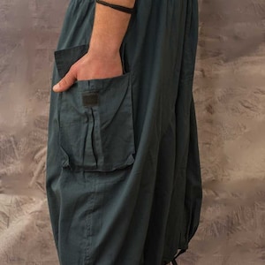side view of Dark grey , loose-fitting trousers with a large cargo pocket on the left side. The trousers have a drawstring waistband and tie closures at the bottom of the legs.