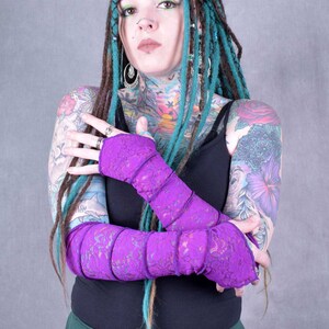 May include: A person with teal dreadlocks and tattoos is wearing a pair of purple lace arm warmers. The arm warmers are folded over and the person is holding their arms in front of their chest.