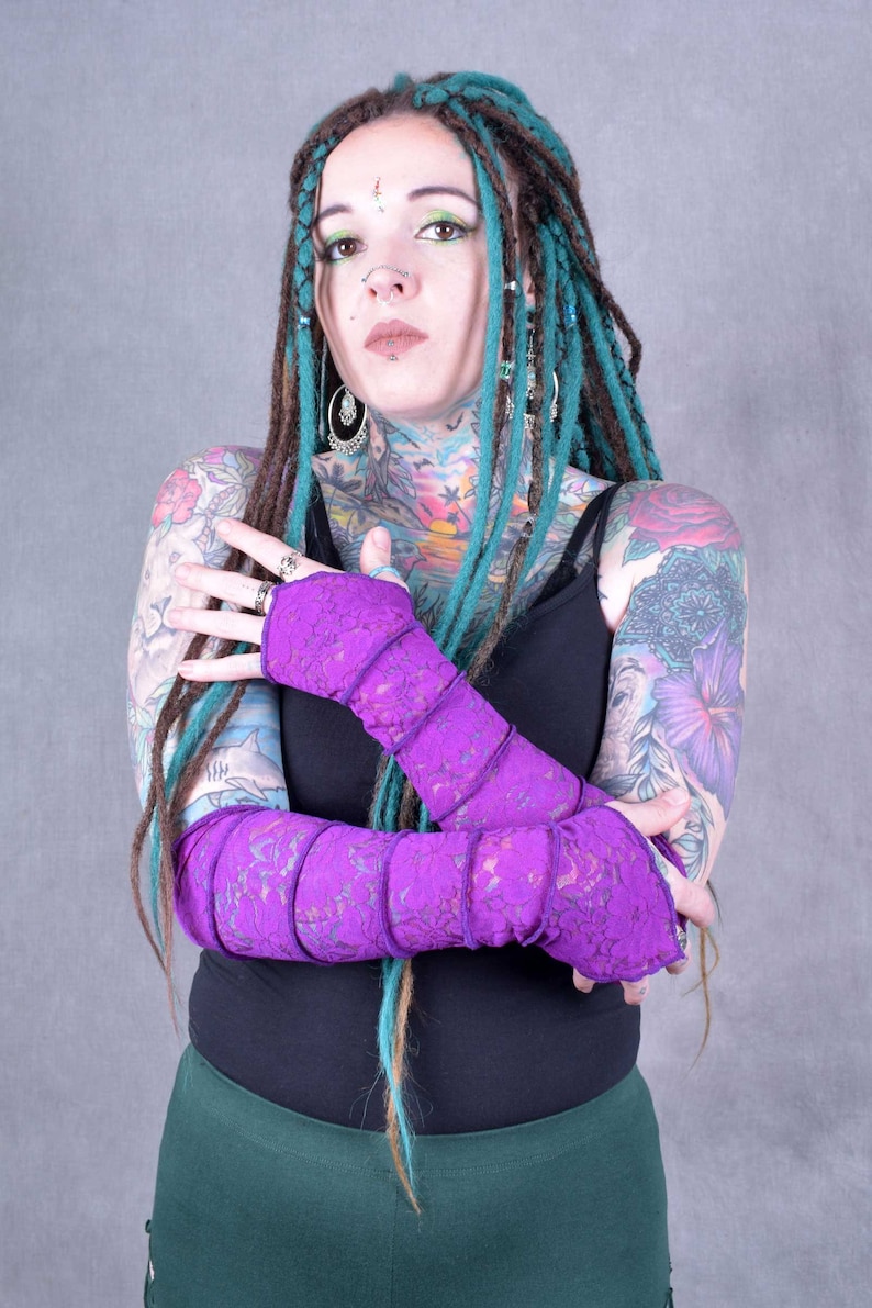 May include: A person with teal dreadlocks and tattoos is wearing a pair of purple lace arm warmers. The arm warmers are folded over and the person is holding their arms in front of their chest.