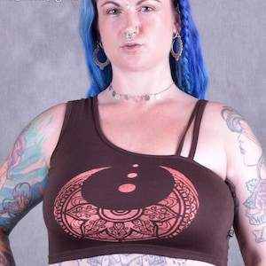 May include: A brown crop top with a pink mandala design and a moon graphic. The top has a one-shoulder design and adjustable straps. The model is wearing the top with a silver necklace and earrings.