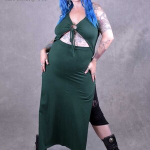 A woman wearing a green maxi dress with a cut-out design.  The dress has a ring detail at the neckline. She is wearing black leggings and black boots. The background is a grey wall. The text "Wobble & Squeak Clothing Co." is visible in the top left corner of the image.