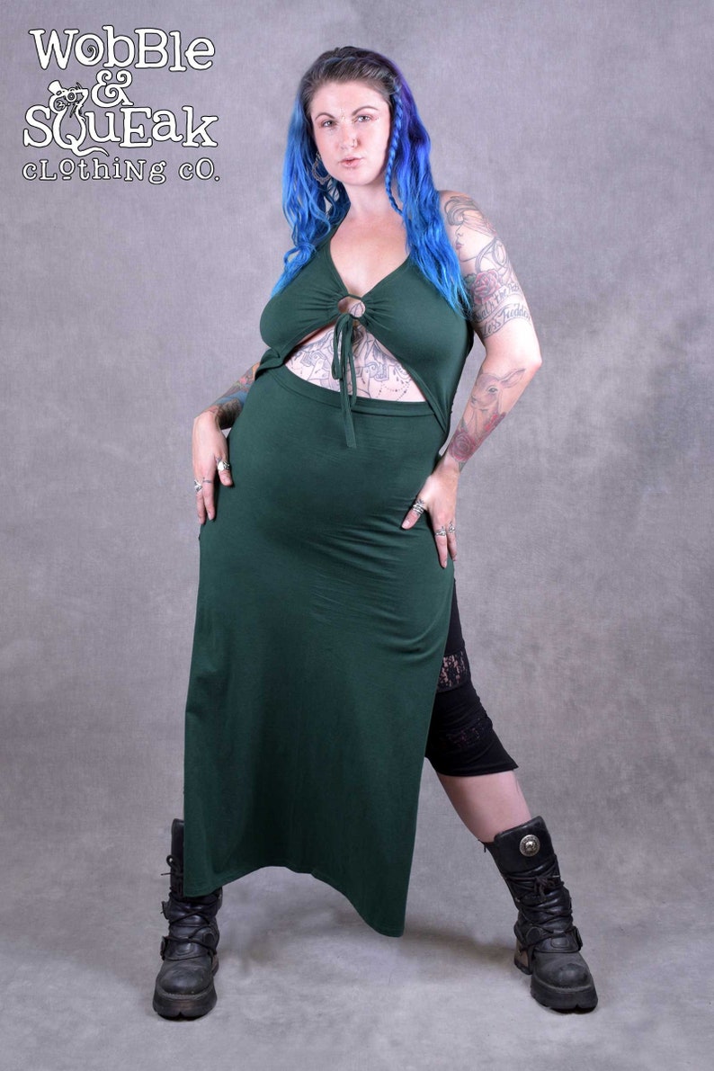 A woman wearing a green maxi dress with a cut-out design.  The dress has a ring detail at the neckline. She is wearing black leggings and black boots. The background is a grey wall. The text "Wobble & Squeak Clothing Co." is visible in the top left corner of the image.