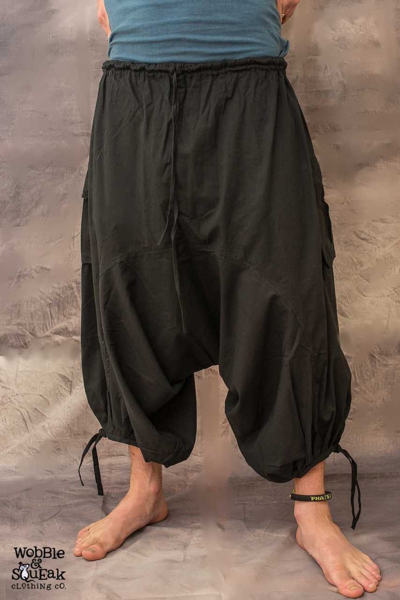 Black cotton trousers with a loose, baggy fit and a drawstring waist. The trousers have two large velcro pockets on the sides and a gathered detail at the ankles.
