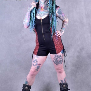 Peut inclure: Une femme avec des dreadlocks bleues et vertes porte une combinaison courte noire et rouge avec une fermeture éclair sur le devant. Elle porte des bottes noires avec des boucles argentées. La combinaison a un motif floral rouge sur les côtés. La femme a des tatouages sur les bras et les jambes.