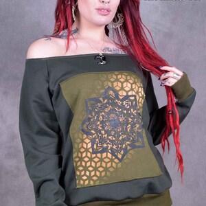 May include: A dark green off-the-shoulder sweatshirt with a gold and black geometric design on the front. The sweatshirt has a ribbed hem and cuffs. The text "Wobble & Squeak clothing co." is printed in white on the top right corner of the image.