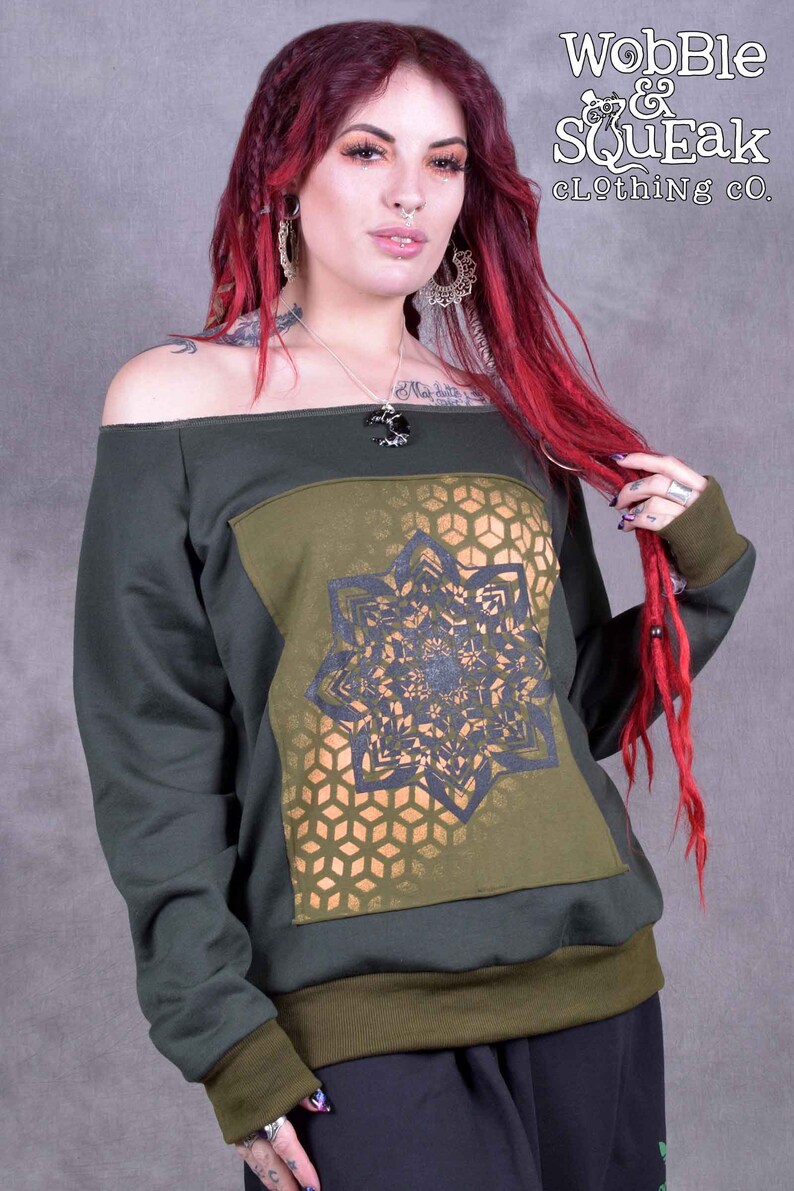 May include: A dark green off-the-shoulder sweatshirt with a gold and black geometric design on the front. The sweatshirt has a ribbed hem and cuffs. The text "Wobble & Squeak clothing co." is printed in white on the top right corner of the image.