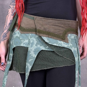 Puede incluir: Una falda de envoltura verde y azul con un estampado floral y un cinturón de tela marrón. La falda está hecha de diferentes capas de tela y tiene un panel de malla. La falda se ata a la cintura y tiene lazos largos que cuelgan a los lados. La falda es de la marca "Wobble & Squeak Clothing Co."