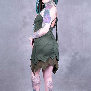 May include: A woman with teal dreadlocks and tattoos wears a green leaf-patterned dress with a lace-up back. The dress has a layered skirt with a jagged hemline.