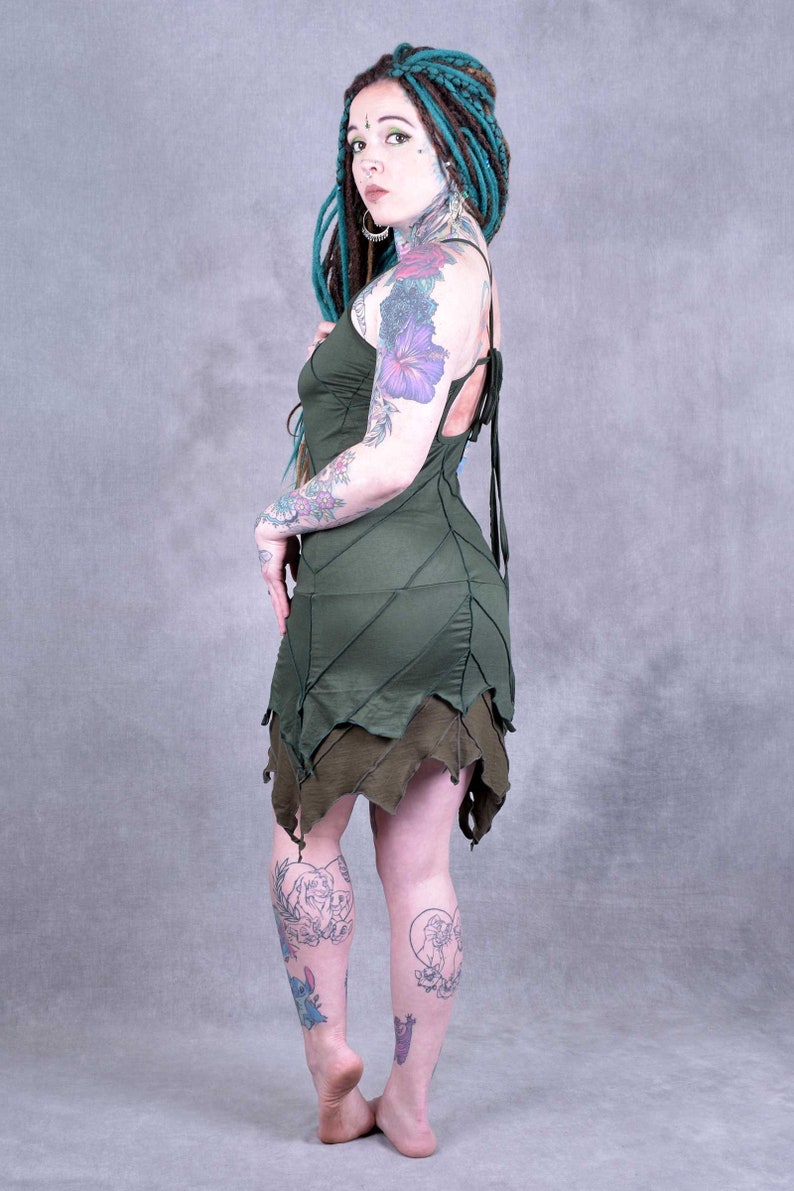 May include: A woman with teal dreadlocks and tattoos wears a green leaf-patterned dress with a lace-up back. The dress has a layered skirt with a jagged hemline.