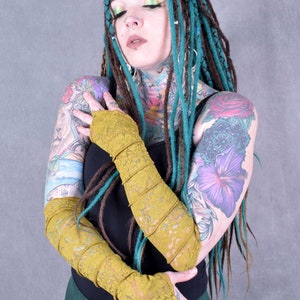May include: A person with teal and brown dreadlocks is wearing a black top and mustard yellow lace fingerless gloves. They have several tattoos on their arms and chest.