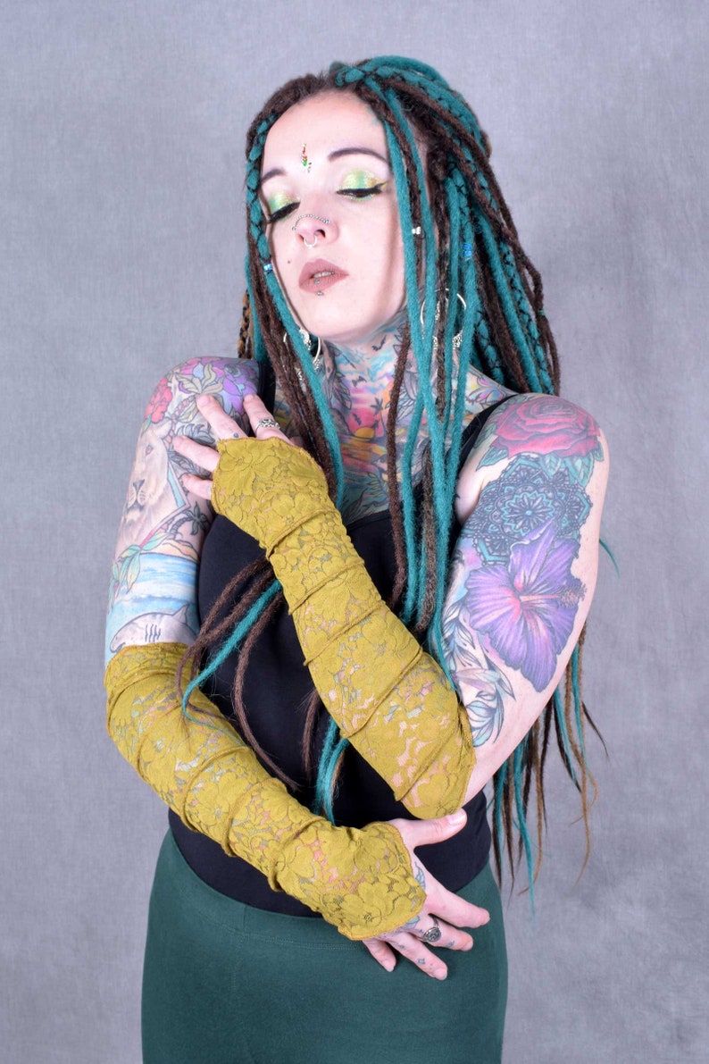 May include: A person with teal and brown dreadlocks is wearing a black top and mustard yellow lace fingerless gloves. They have several tattoos on their arms and chest.