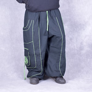 May include: Dark grey baggy trousers with neon green trim and accents. The trousers feature large pockets and a drawstring waist. The design includes a spiral detail on one pocket. The trousers are worn with dark shoes and a black top.