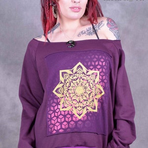 May include: Purple off-the-shoulder sweatshirt with a gold and pink mandala design. The sweatshirt has a purple trim around the neckline and cuffs. The text "Wobble & Squeak Clothing Co." is printed on the top right corner of the image.