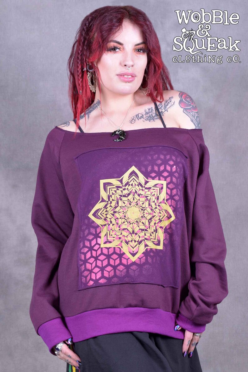 May include: Purple off-the-shoulder sweatshirt with a gold and pink mandala design. The sweatshirt has a purple trim around the neckline and cuffs. The text "Wobble & Squeak Clothing Co." is printed on the top right corner of the image.