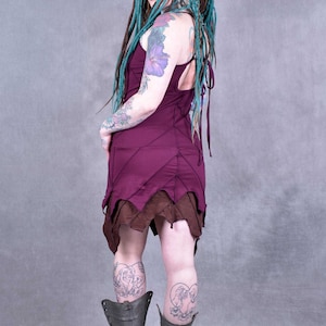 May include: A woman wearing a burgundy dress with brown trim and green dreadlocks. She is wearing black boots and has tattoos on her arms and legs.
