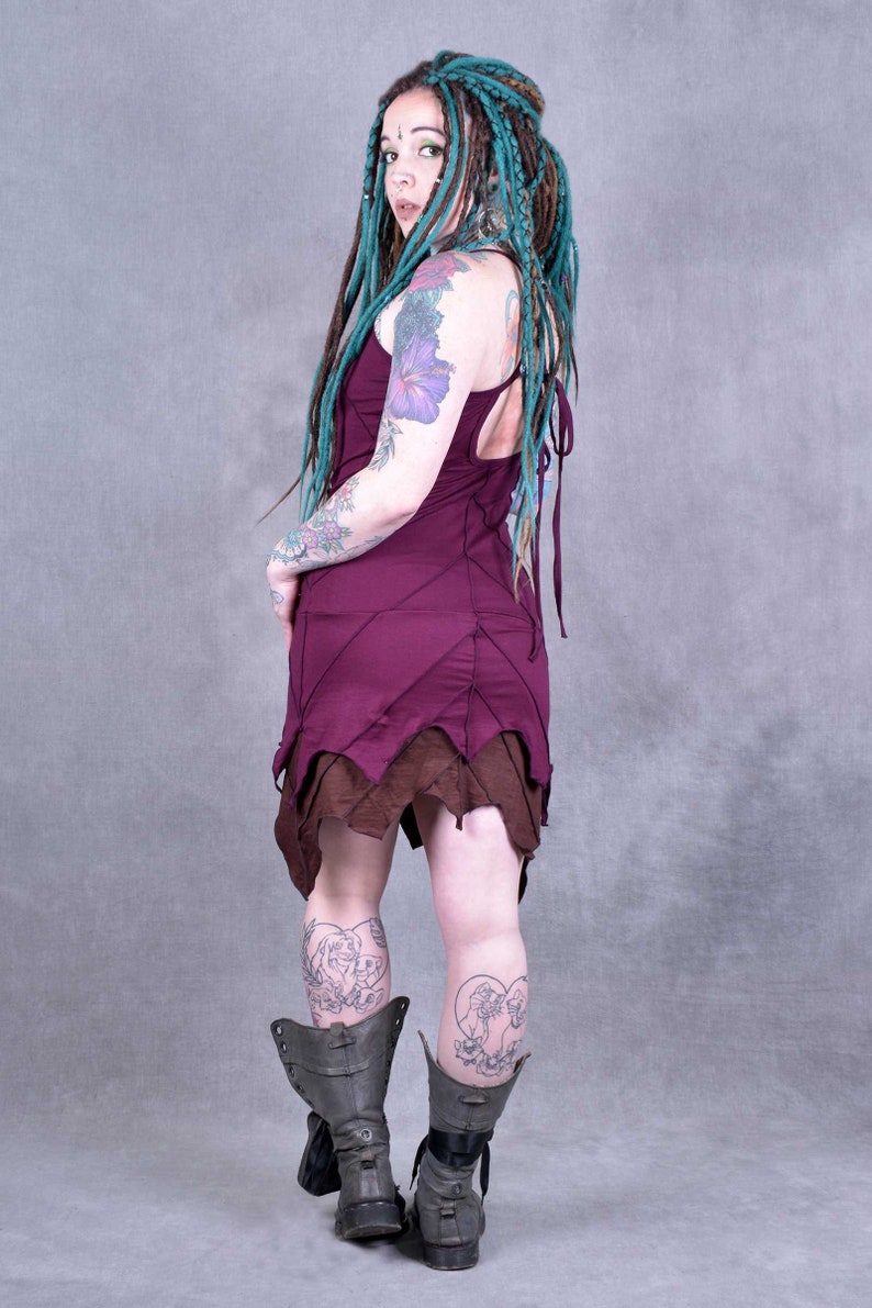 May include: A woman wearing a burgundy dress with brown trim and green dreadlocks. She is wearing black boots and has tattoos on her arms and legs.