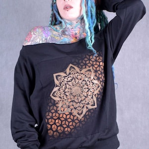 Puede incluir: Sudadera negra de hombros descubiertos con un diseño de mandala geométrico dorado. La sudadera tiene mangas largas y un corte holgado.