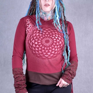 Könnte beinhalten: Ein langärmliges Hemd in Rotbraun mit einem braunen Band an der Taille. Das Hemd hat ein geometrisches Muster auf der Vorderseite und Spitzenmanschetten. Das Model trägt eine schwarze Hose und hat blaue und grüne Dreadlocks.