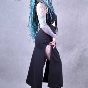 A woman with blue and green dreadlocks is wearing a black maxi dress with a side slit and a tie at the top. The dress is made of a soft, flowing fabric. She is wearing black platform boots with buckles. The background is a light grey.