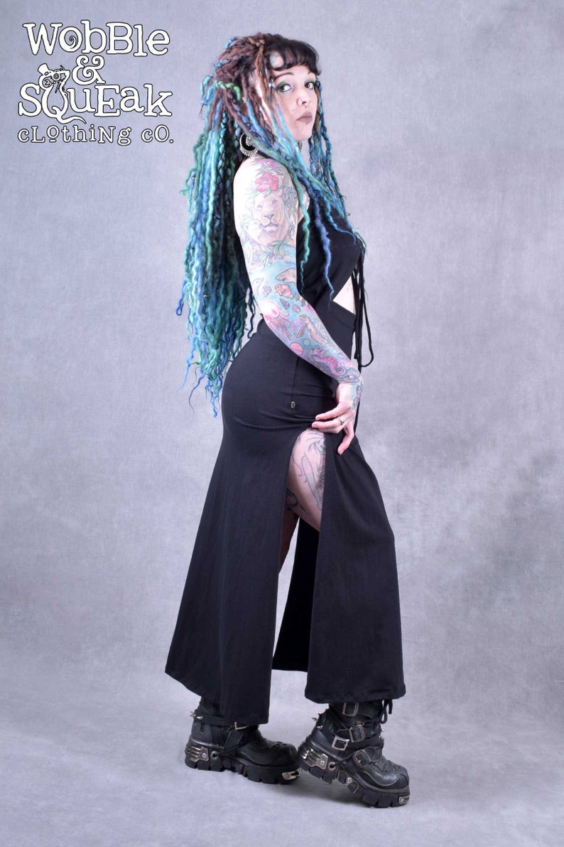 A woman with blue and green dreadlocks is wearing a black maxi dress with a side slit and a tie at the top. The dress is made of a soft, flowing fabric. She is wearing black platform boots with buckles. The background is a light grey.
