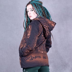 Puede incluir: Una chaqueta negra con capucha con un patrón geométrico marrón y el texto "RESONATE" en la espalda. La chaqueta tiene una cremallera y una capucha con cordón.