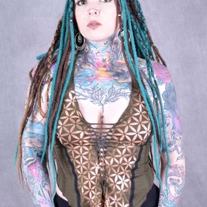Könnte beinhalten: Eine Frau mit türkisfarbenen und braunen Dreadlocks trägt ein grünes Halstuch mit einem braunen geometrischen Muster. Sie hat mehrere Tattoos auf ihren Armen und ihrer Brust.