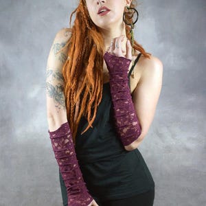 May include: A woman wearing a black tank top and black pants with a pair of long, burgundy lace fingerless gloves. The gloves are gathered at the wrist and have a delicate lace pattern. The woman has long, red dreadlocks and is wearing makeup. The image includes the text "Wobble & SouEak Clothing Co."