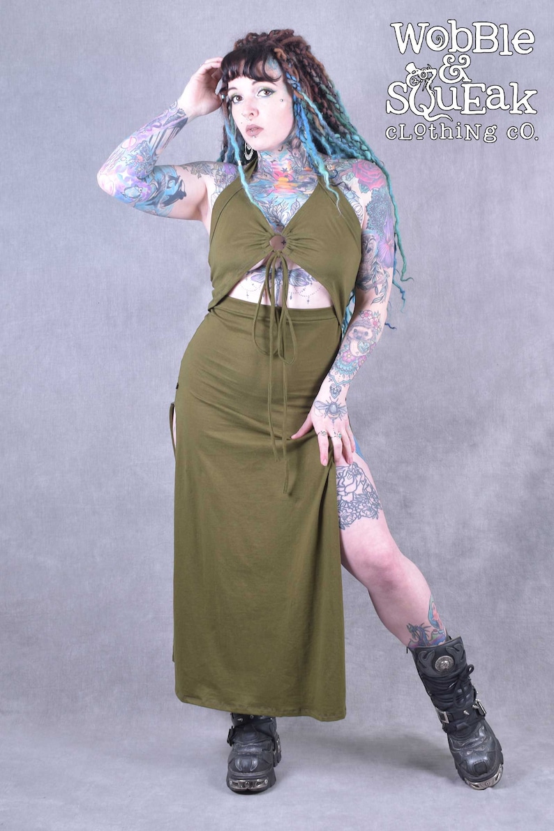 A long, army green maxi dress with a halter neckline and a tie at the waist. The dress has a side slit and a button detail at the chest. The model is wearing the dress with black boots.