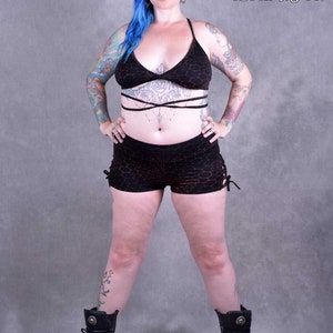 Könnte beinhalten: Eine Frau trägt ein schwarzes und orangefarbenes gemustertes Crop-Top und eine passende Shorts. Sie hat blaues Haar und Tattoos auf ihren Armen. Die Shorts haben einen seitlichen Knoten. Sie trägt schwarze Stiefel. Das Bild enthält den Text "Wobble & Squeak clothing co."
