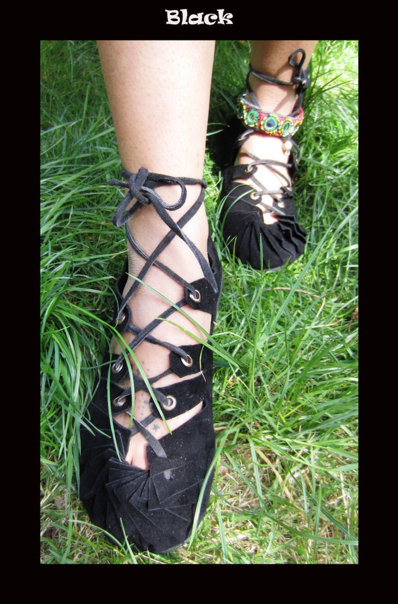 Black leather sandals with laces tied up the leg. The sandals have a unique design with a pointed toe and a thick sole. The sandals are worn by a person standing in a grassy field.