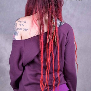 May include: A purple off-the-shoulder sweatshirt with long sleeves and a purple waistband. The model has red and blonde dreadlocks and a tattoo on their left shoulder that says "Take a ride on the Wild Side".