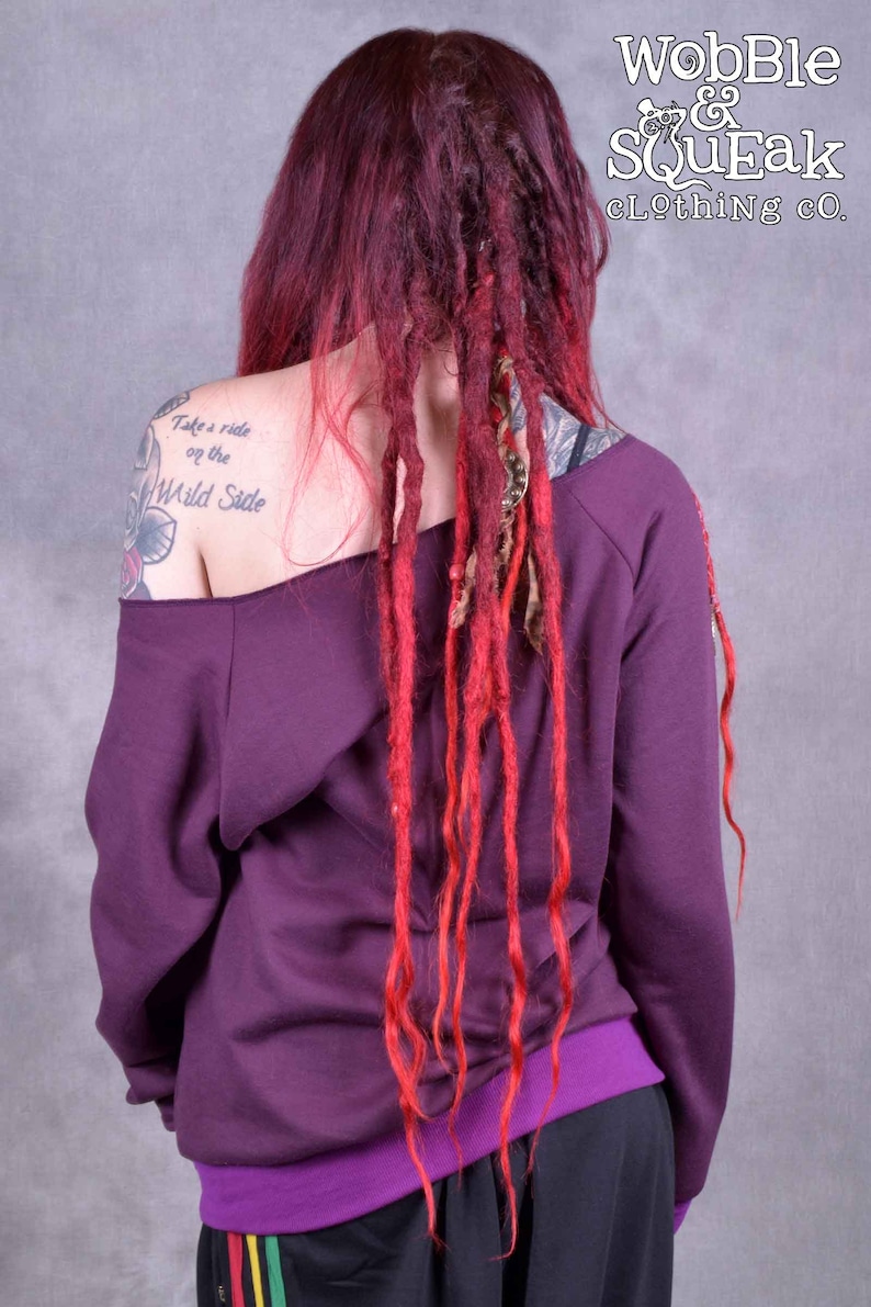 May include: A purple off-the-shoulder sweatshirt with long sleeves and a purple waistband. The model has red and blonde dreadlocks and a tattoo on their left shoulder that says "Take a ride on the Wild Side".