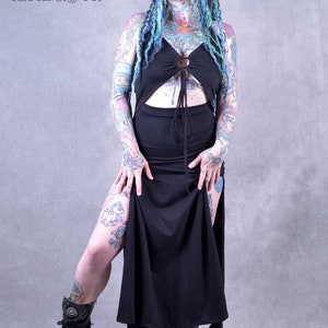 A black maxi dress with a cut-out design showing the tummy area and a tie at the waist. The dress has a long slit on the side and is worn with black boots. The model has blue and green dreadlocks and tattoos.