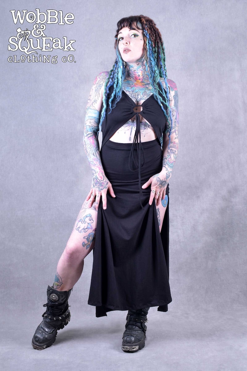 A black maxi dress with a cut-out design showing the tummy area and a tie at the waist. The dress has a long slit on the side and is worn with black boots. The model has blue and green dreadlocks and tattoos.