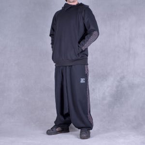May include: A black hooded sweatshirt with a zipped collar and patterned elbow patches. The sweatshirt is paired with black trousers with a patterned stripe down the side. The trousers have a drawstring waist and a loose fit.