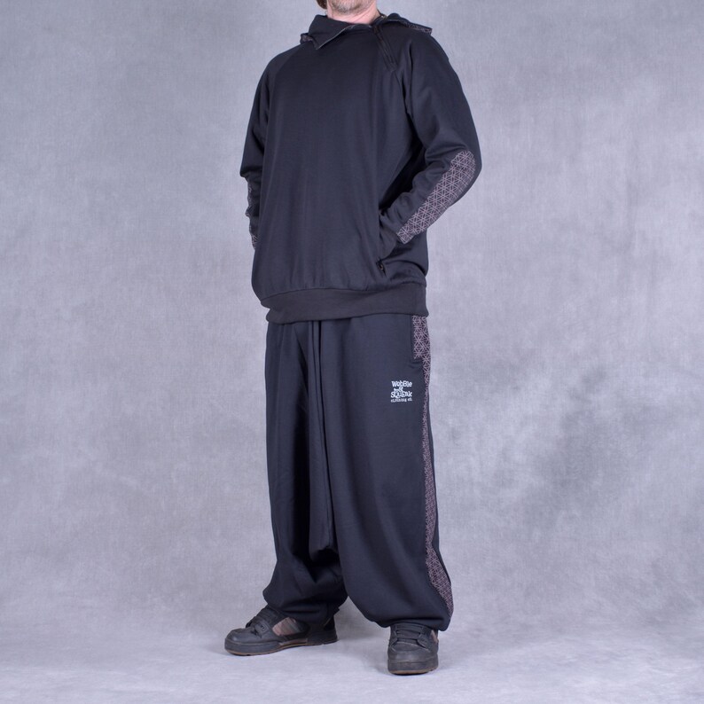 May include: A black hooded sweatshirt with a zipped collar and patterned elbow patches. The sweatshirt is paired with black trousers with a patterned stripe down the side. The trousers have a drawstring waist and a loose fit.