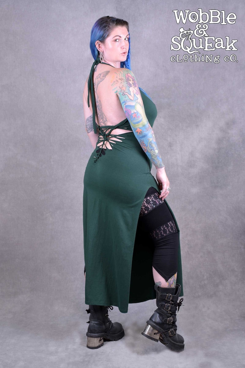A dark green maxi dress with a lace-up back and a side slit. The dress is made from a soft, stretchy fabric and has a relaxed fit. The model is wearing the dress with black leggings and boots. it also has a knotted detail at the back