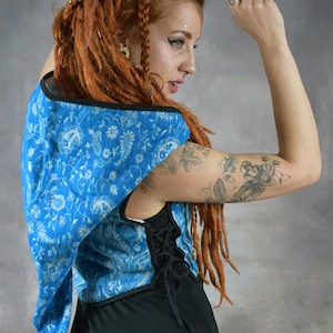 Puede incluir: Una mujer con largas rastas rojas lleva un chaleco con capucha azul y blanco con estampado y ribetes negros. El chaleco tiene una espalda con cordones y se lleva sobre una camiseta negra de tirantes.