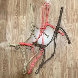 May include: Three rope halters in beige, neon orange, and dark brown. Each halter is constructed from thick, braided cord featuring adjustable knots and a circular metal ring at the chin area. The design includes multiple loops and secure knots.