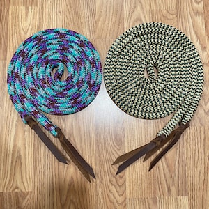 May include: Two coiled horse reins. One is turquoise, purple, and burgundy, the other is black and tan. Both have brown leather ends. The reins are on a wooden surface.