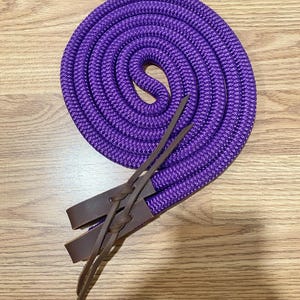 May include: A coiled purple rope with brown leather detailing. The rope is tightly woven and appears robust. The leather straps are attached to the rope with intricate knots. This item is likely used for equestrian activities.